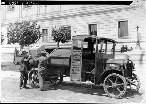 Two men dumping waste into the back of an old early 20th century Street Cleaning truck in front of San Francisco City Hall.